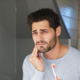 Wincing man holding his cheek in one hand and a toothbrush in the other